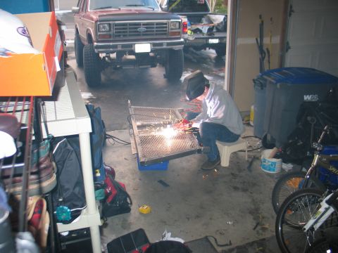 1984 Ford Bronco - Person welding a metal frame in a garage with a Ford Bronco and another truck visible.