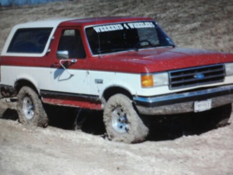 1989 Ford Bronco - This red and white Ford Bronco appears to be from the late 1980s, possibly a 1987 model, stuck in a muddy off-road environment.