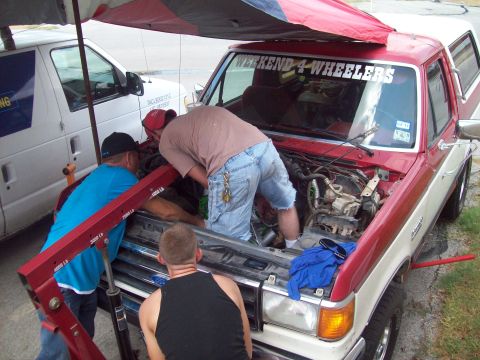 1989 Ford Bronco - Mechanics work on the engine of a red and white 1989 Ford Bronco, with a white van nearby.