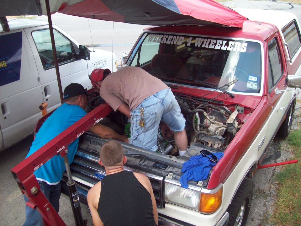 1989 Ford Bronco with engine out, people working on it. White van in background