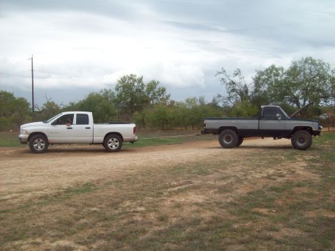 1989 Ford Bronco - A white pickup truck tows a modified gray pickup truck across a dirt lot under a cloudy sky.