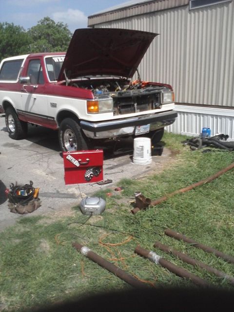 1989 Ford Bronco - A two-tone red and white Ford Bronco is parked with its hood open, surrounded by tools and pipes.