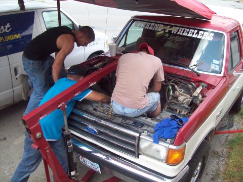 1989 Ford Bronco - Mechanics work on the engine of a red and white Ford Bronco, with a white van nearby.