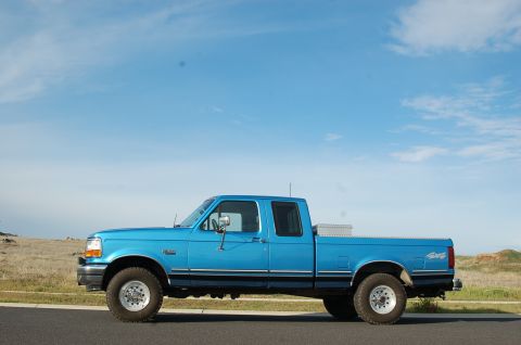 1992 Ford F-150 - A blue 1992 Ford F-150 truck is parked on the side of the road under a clear blue sky.