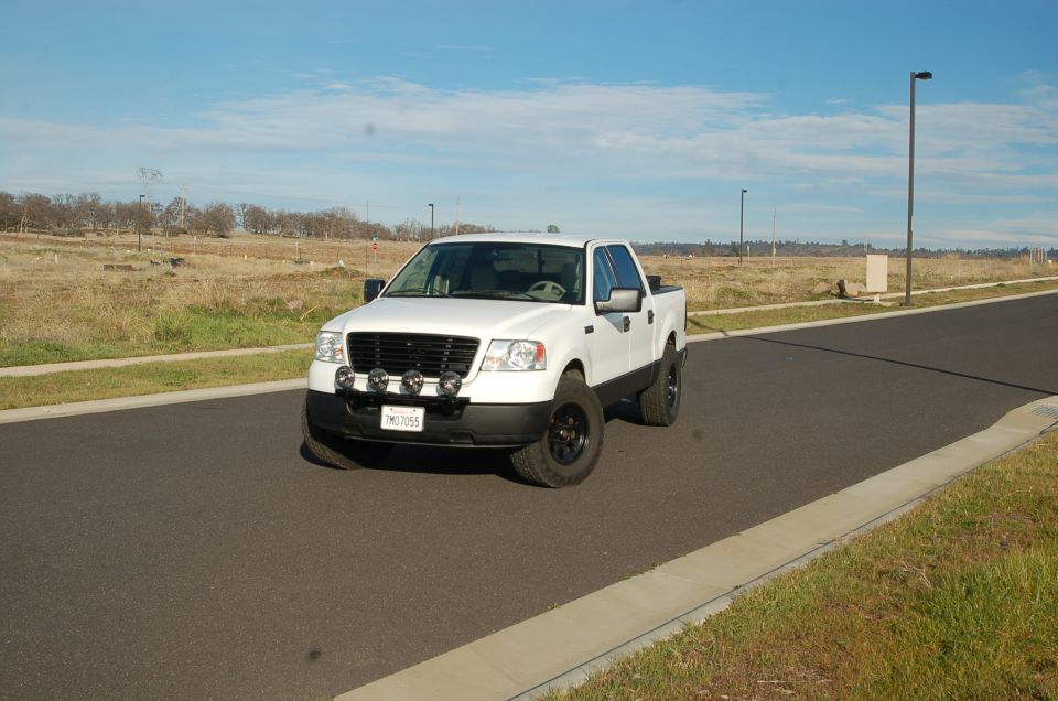 White Ford F-150 pickup truck with aftermarket lights and black wheels, parked on a road