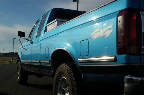 1992 Ford F-150 - A blue 1992 Ford F-150 pickup truck is viewed from the 3/4 front angle, showing its 4x4 decal and a toolbox in the bed.