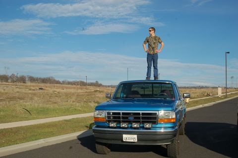 1992 Ford F-150 - A person stands atop a blue 1992 Ford F-150 pickup truck parked on the side of a road under a partly cloudy sky.