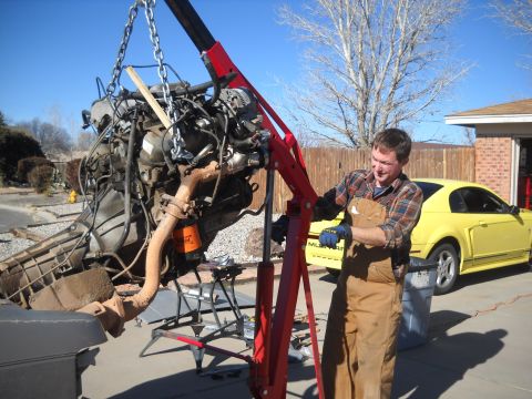 1992 Ford Explorer - A man uses an engine hoist to remove a vehicle's engine. A yellow car is parked nearby.