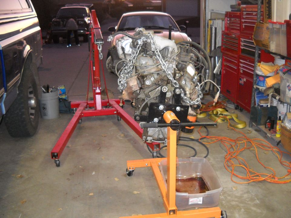 Engine on an orange engine stand, with a red engine hoist nearby. A pickup truck is partially visible on the left