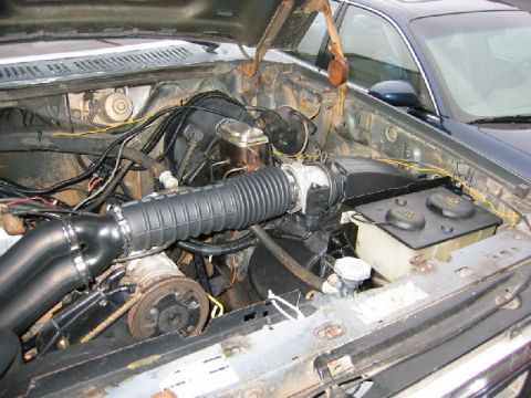 1986 Ford Bronco - The engine bay of a vehicle, showing various mechanical parts and wiring, with another vehicle visible in the background.