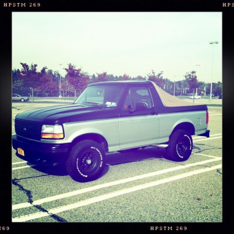 1992 Ford Bronco - A two-tone 1992 Ford Bronco with a tan soft top is parked in a lot under a clear sky.