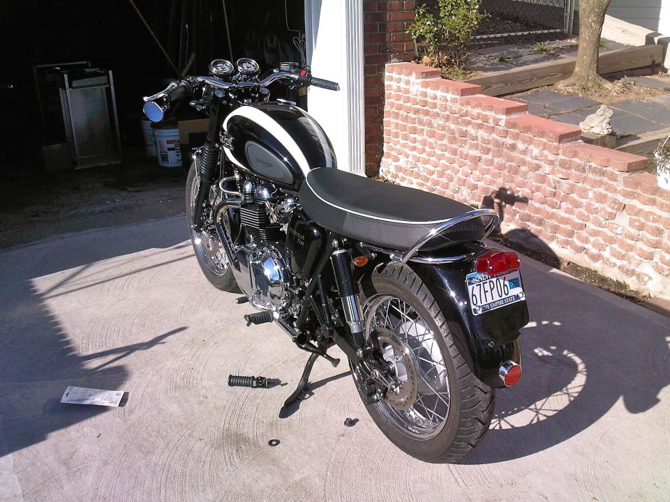 Black Triumph motorcycle, rear 3/4 view, white tank accent, NY plate, parked on driveway by garage