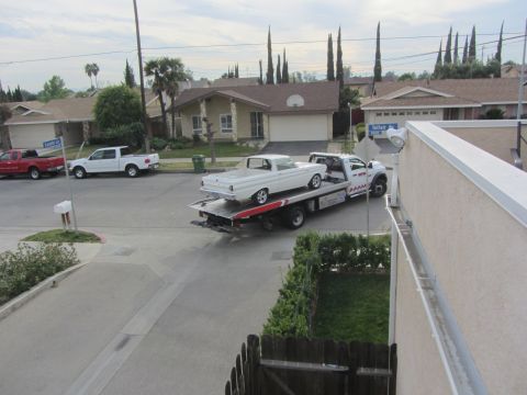 1964 Ford Ranchero - A white pickup truck is being towed on a flatbed truck down a suburban street.