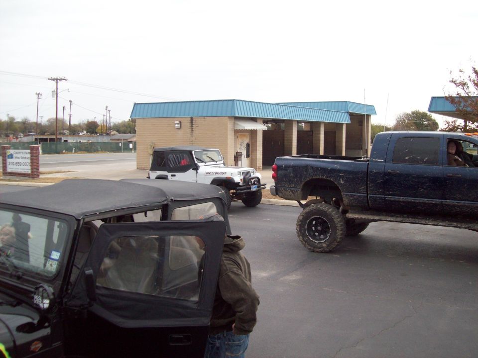 Three lifted off-road vehicles parked near a building with a blue roof. One is a black Jeep in the foreground, another is a white Jeep, and the third is a dark blue pickup truck