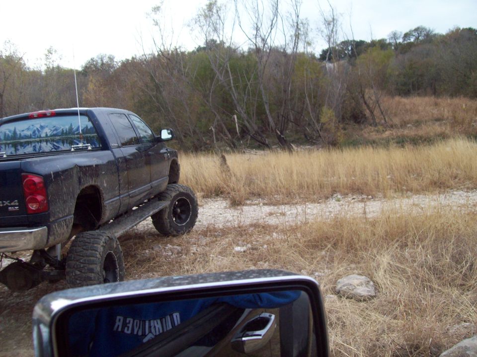 Lifted black pickup truck with large tires parked on a dirt path next to dry grass and trees. Side mirror reflects a blue shirt