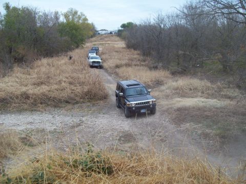 1989 Ford Bronco - A convoy of SUVs, led by a black Hummer H3, travels down a dusty, unpaved road through a dry, wooded area.