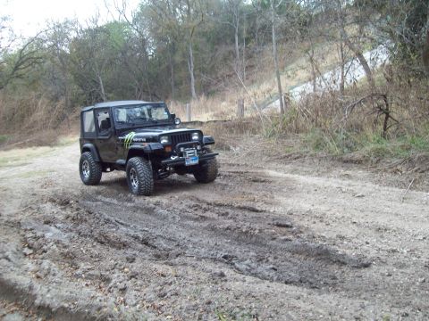 1989 Ford Bronco - A black Jeep Wrangler navigates a muddy off-road path surrounded by trees and dry brush.