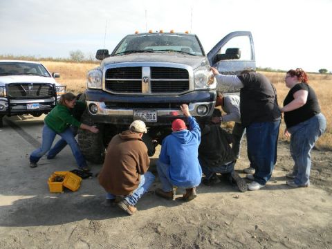 1989 Ford Bronco - A group of people gather around a dark blue Dodge Ram truck, appearing to work on its front end, with another white truck visible behind it.