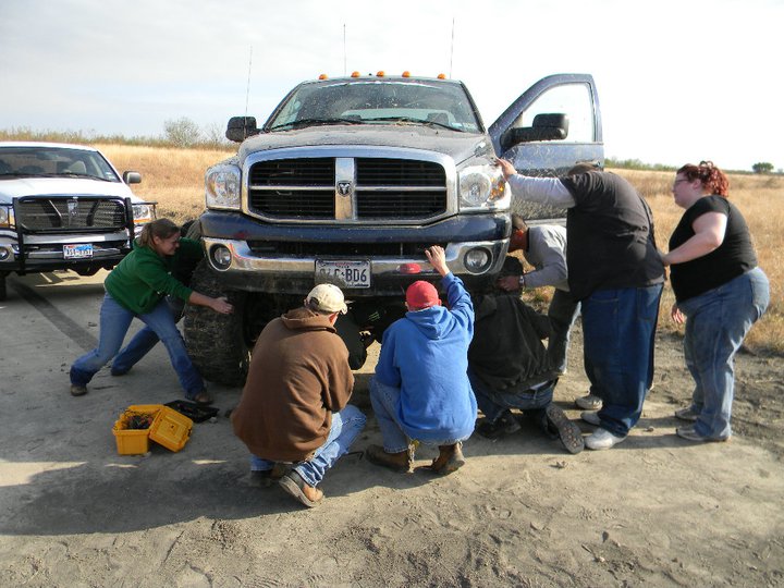 Front view of dark blue Dodge Ram truck with people working on it; white truck in background