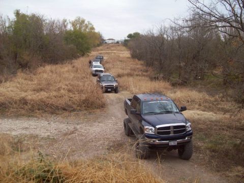 1989 Ford Bronco - Multiple SUVs and trucks are seen driving in a convoy on a dirt path.