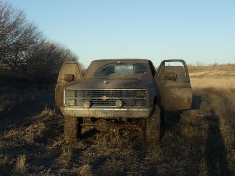 1989 Ford Bronco - This heavily mud-caked Chevrolet C/K pickup truck, likely from the mid-1980s, is parked in a grassy field with its doors ajar.