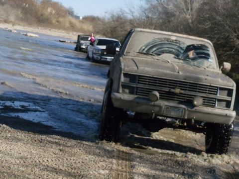 1989 Ford Bronco - Several muddy pickup trucks navigate through a shallow body of water.
