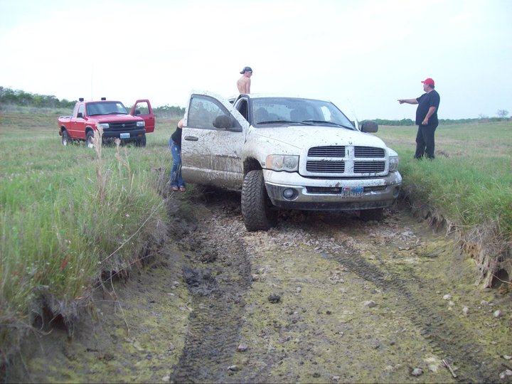 White pickup truck stuck in mud with people around. Red pickup truck in background
