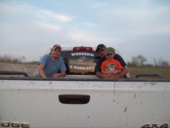 Three people, including a child, sit in the bed of a white pickup truck. A sticker on the back window reads 'WEEKEND 4 WHEELERS'