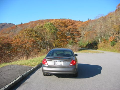 2003 Ford Taurus - A gray 2003 Ford Taurus is parked on a road with colorful autumn trees on a hill under a clear blue sky.