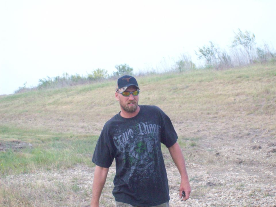 Man wearing a black t-shirt with 'Grave Digger' graphic and a Texas Longhorns cap stands in a grassy field