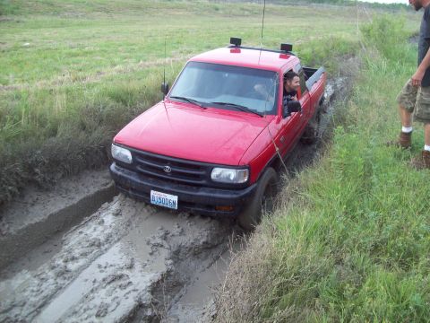 1989 Ford Bronco - A red pickup truck navigates a muddy path, with a person in the driver's seat and another standing nearby.