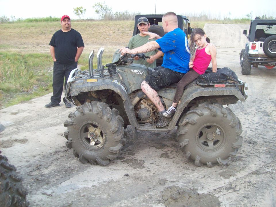 Muddy ATV with two adults and a child riding, two other people standing nearby, and a Jeep in the background