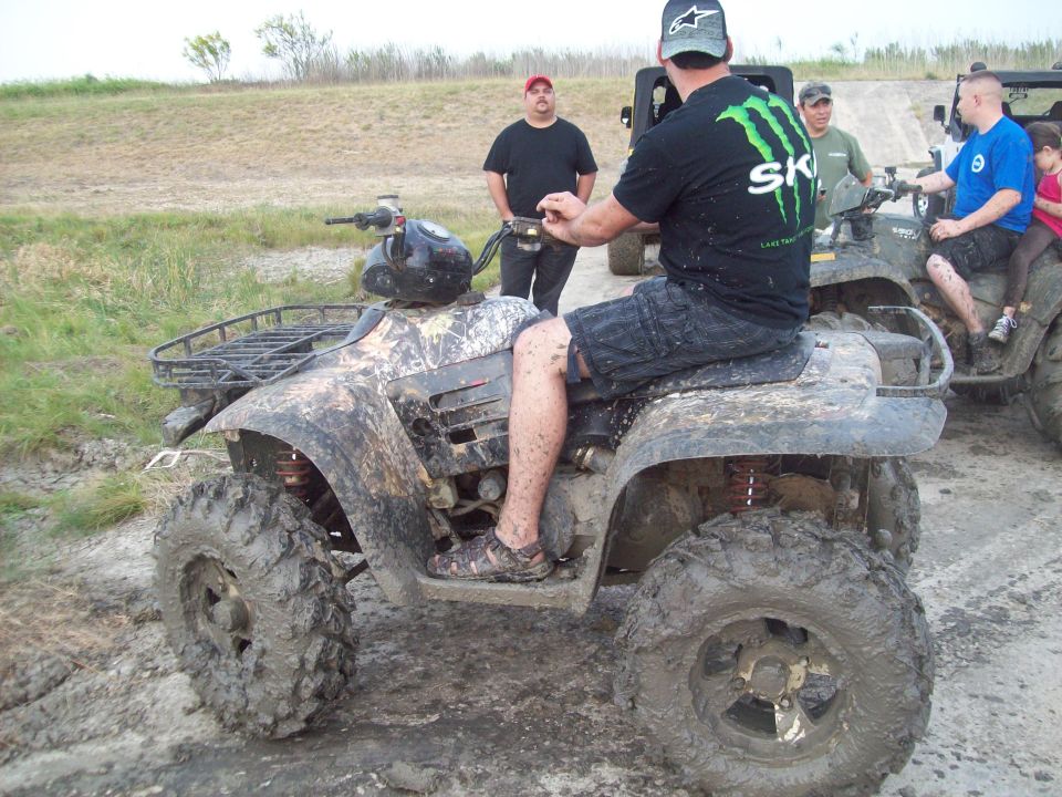 Mud-covered ATV with camouflage pattern, side view. Rider in black shorts and Monster Energy shirt