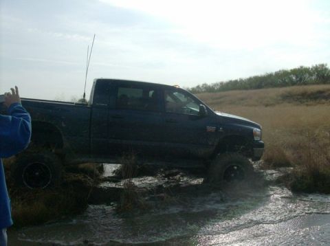 1989 Ford Bronco - A dark blue pickup truck navigates through a muddy, watery terrain, kicking up spray as it moves.