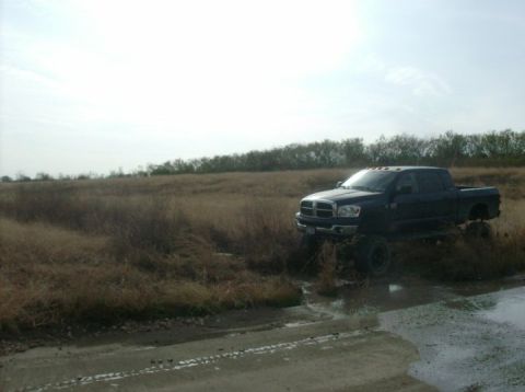 1989 Ford Bronco - Black Dodge Ram pickup truck with lifted suspension driving through a muddy field.
