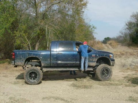 1989 Ford Bronco - A lifted blue pickup truck is parked on a dirt road with trees in the background. Two people are visible near the driver's side door.