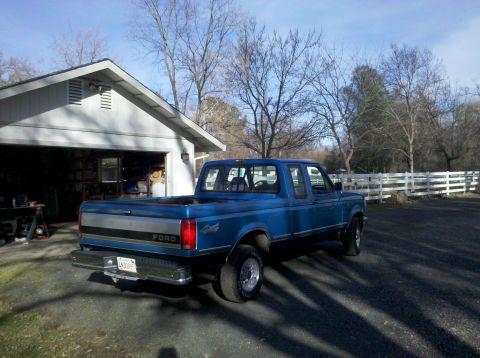 1992 Ford F-150 - A blue 1992 Ford F-150 pickup truck is seen from the rear, parked in front of a garage with a white fence in the background.