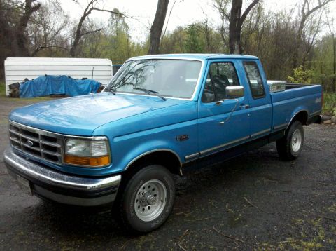 1992 Ford F-150 - A blue 1992 Ford F-150 pickup truck is parked in a driveway with trees and a shed in the background.