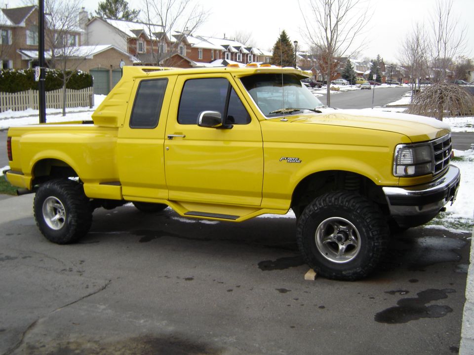 Yellow Ford F-150 pickup truck with custom bed cap, lifted suspension, and aftermarket wheels, parked on a residential street