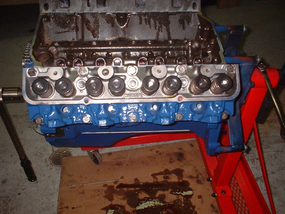 Engine block with blue painted intake manifold and exposed valve train components. The engine is on a red stand