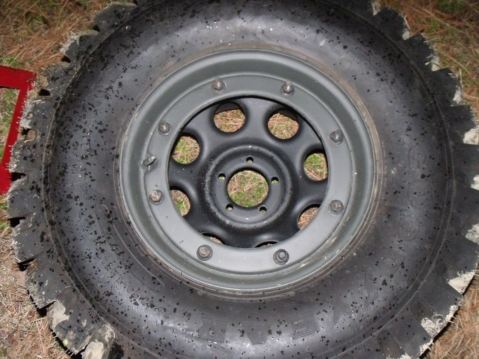 Close-up of a large, knobby off-road tire mounted on a black wheel. Some mud is visible on the tire