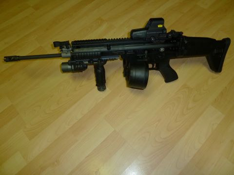 1992 Ford Mustang - FN SCAR-H rifle equipped with a drum magazine and optical sight rests on a wooden floor.