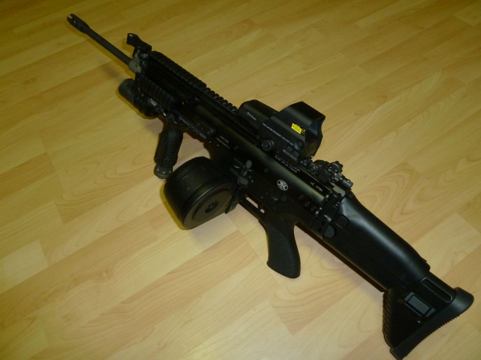 Black assault rifle with drum magazine, red dot sight, and foregrip on a wooden floor