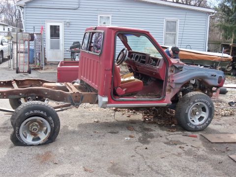 1987 Ford Ranger - A red pickup truck chassis sits in a yard, missing its front end and bed, with a flat front tire.