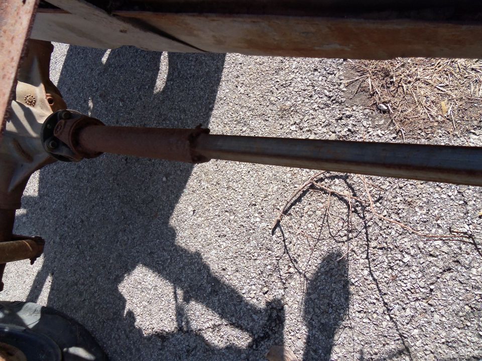 Close-up of a rusty driveshaft connected to a differential, with gravel and asphalt visible