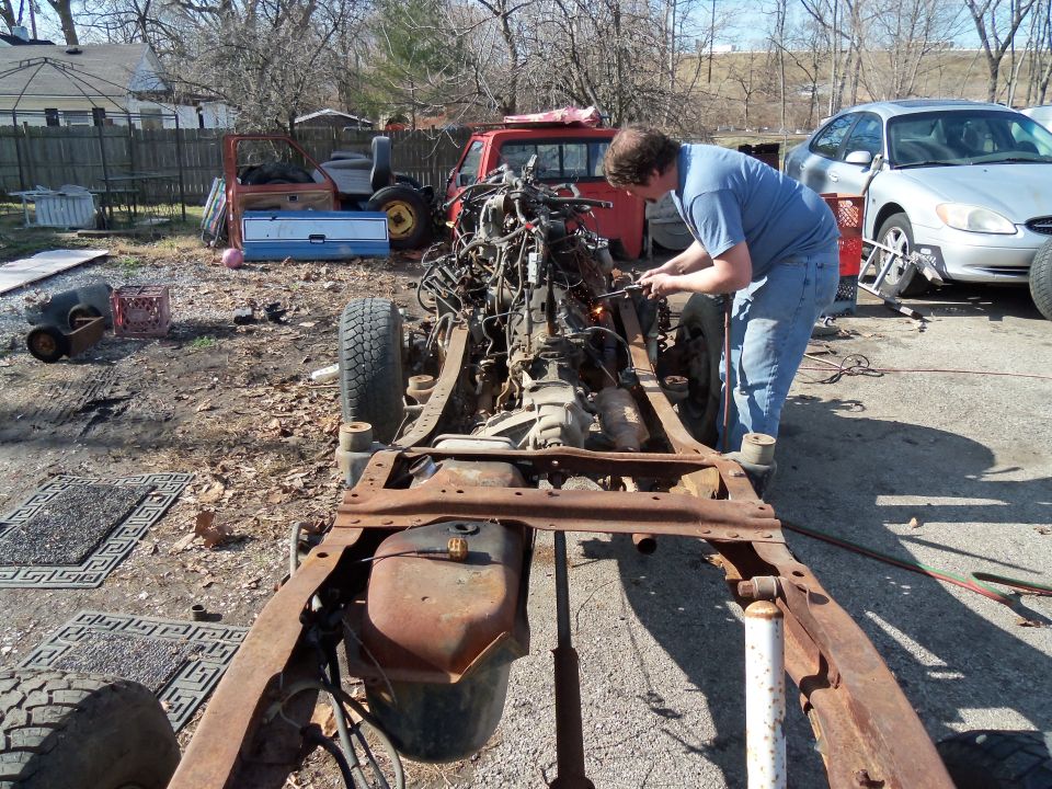 Rusty truck frame with engine and transmission, person welding
