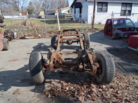 1987 Ford Ranger - A heavily rusted truck chassis sits outdoors with a red truck cab visible in the background.