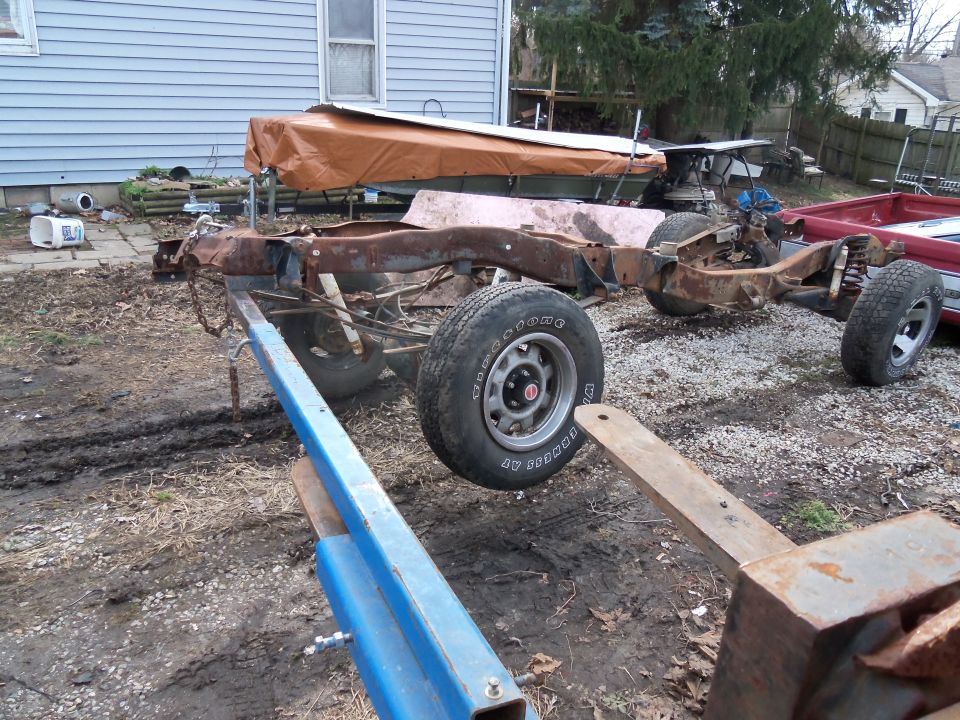 Rusted truck frame and suspension with wheels, partially supported by blue metal equipment