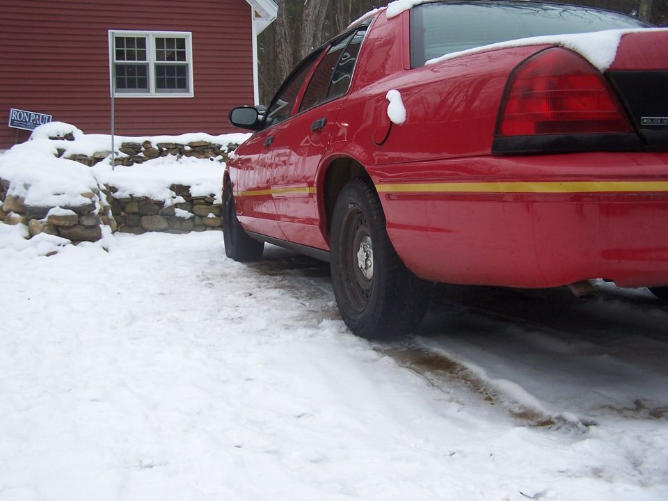 Red Crown Victoria with yellow stripe and Police Interceptor badge parked in snow beside a red house