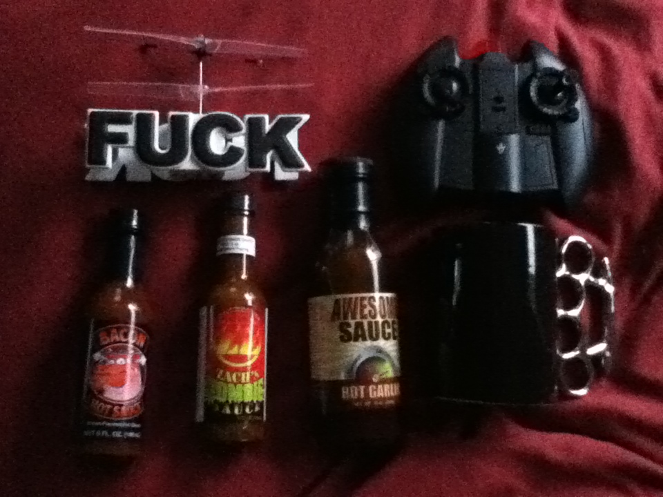 Remote control helicopter with 'FUCK' sign, three bottles of hot sauce, a black mug, and a black remote control